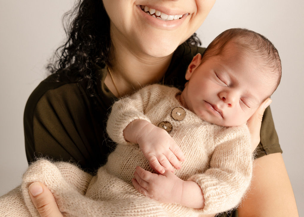 A newborn baby sleeps in a cream knit onesie in mom's arms