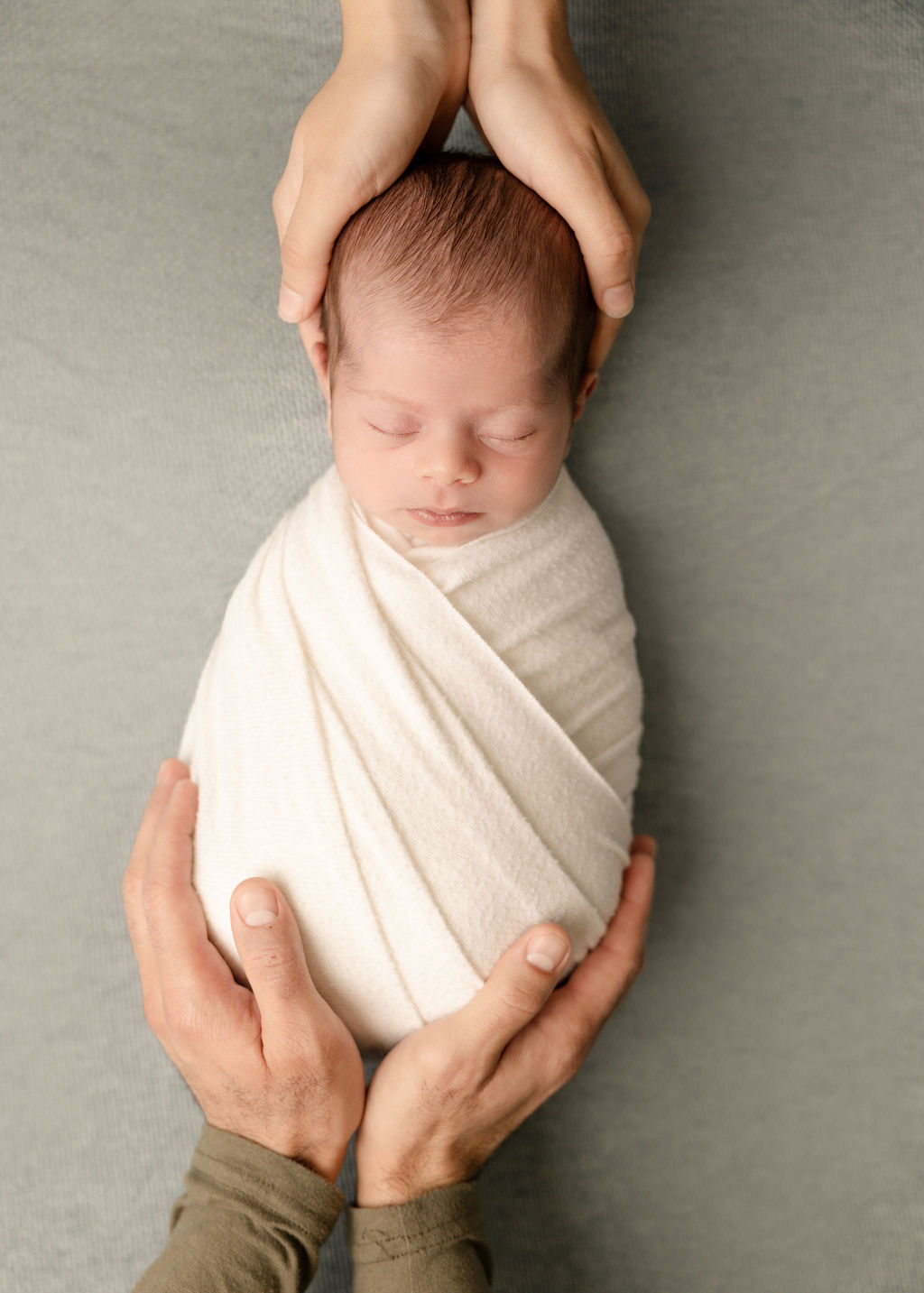 A newborn baby sleeps in an egg swaddle while mom and dad hold its head and feet