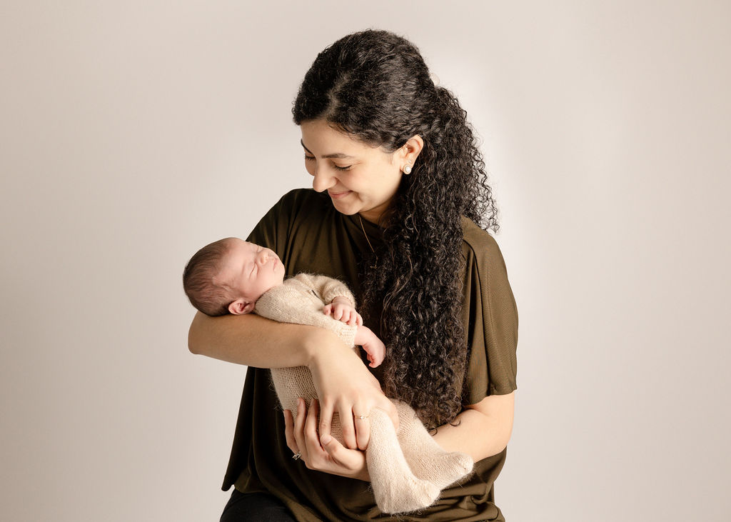 A happy mom smiles down at her sleeping newborn baby in her arms while standing in a studio after meeting a long beach pediatric dentist