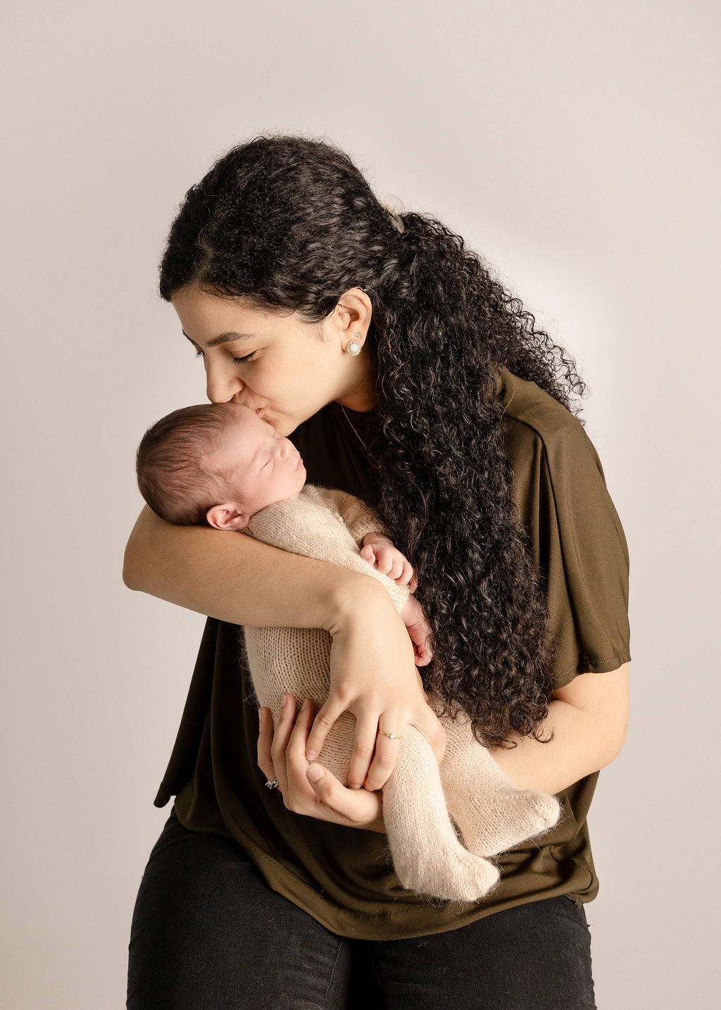 A happy new mom kisses her sleeping newborn while standing in a studio after finding a long beach pediatric dentist