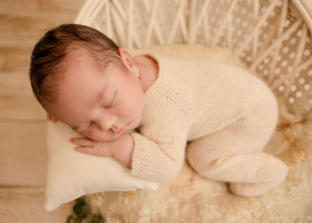 A sleeping newborn baby in a white knit onesie sleeps in a wicker basket on a pillow