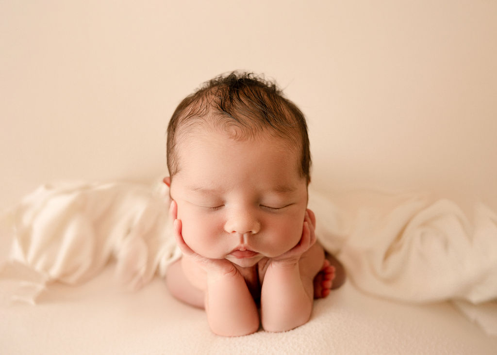 A newborn baby rests its head in its hands while sleeping on a white bed