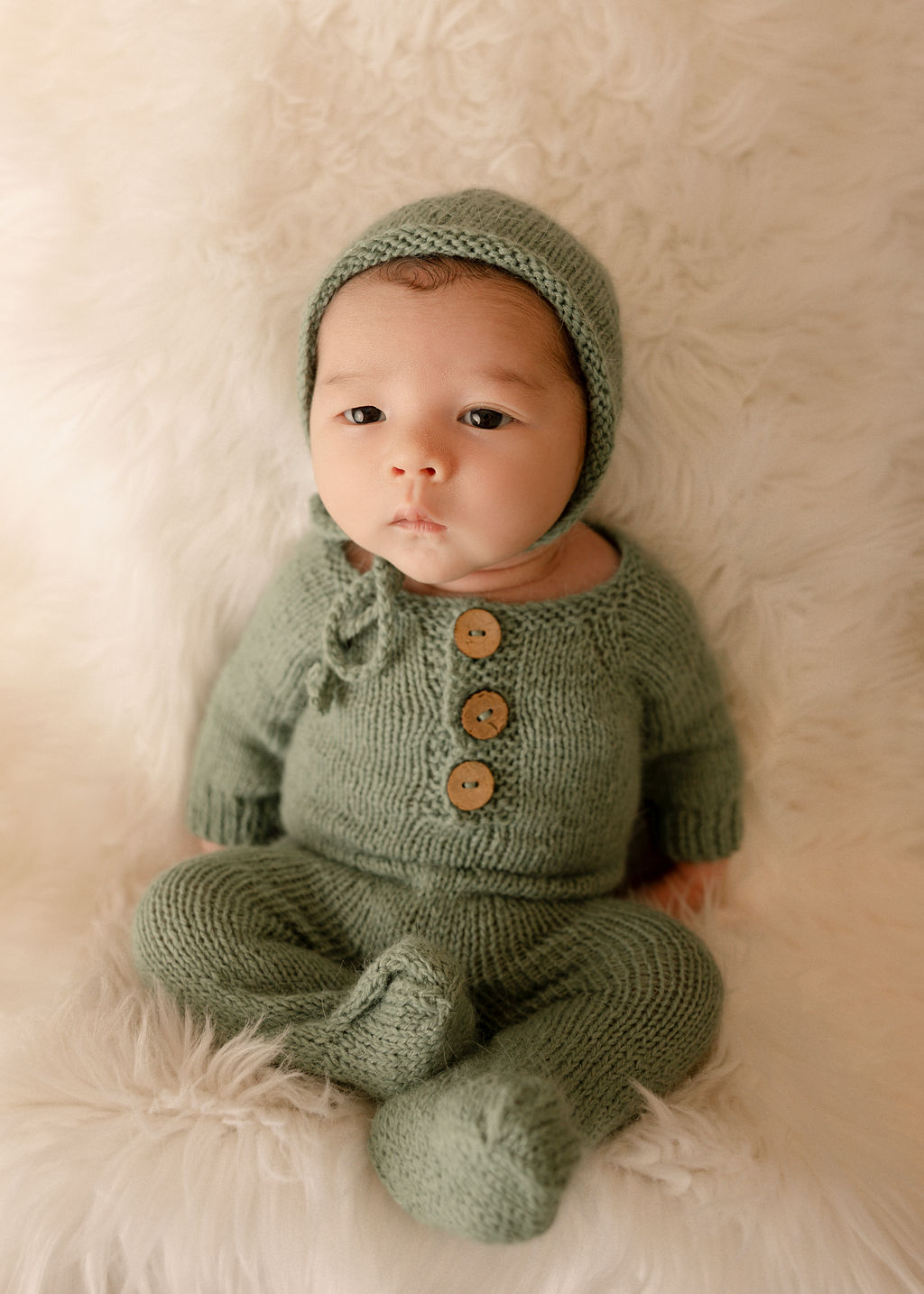 A newborn baby lays on a fur blanket in a green knit onesie and matching bonnet after meeting with long beach pediatricians