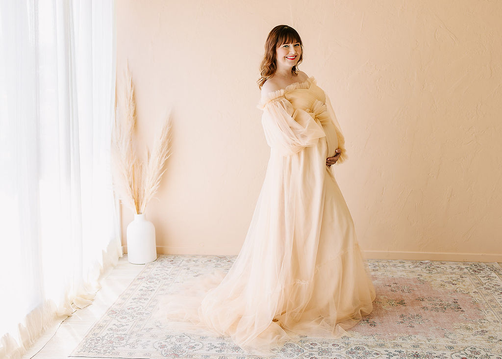 A mom to be stands on a rug in a studio smiling while holding her bump