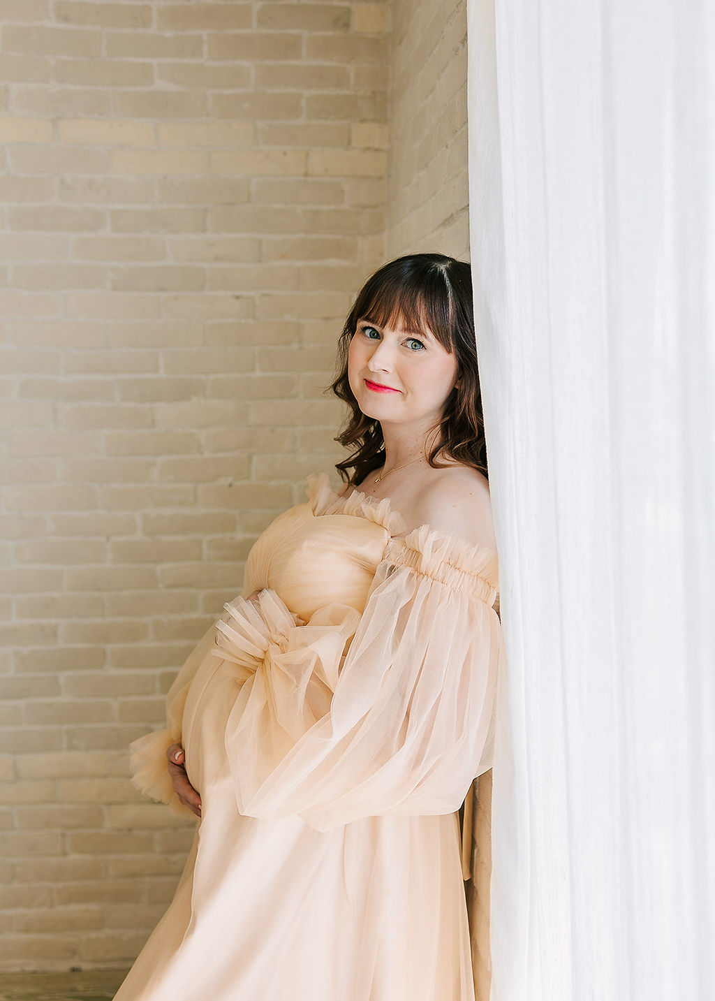 A pregnant woman leans on a brick wall in a studio in a cream maternity gown