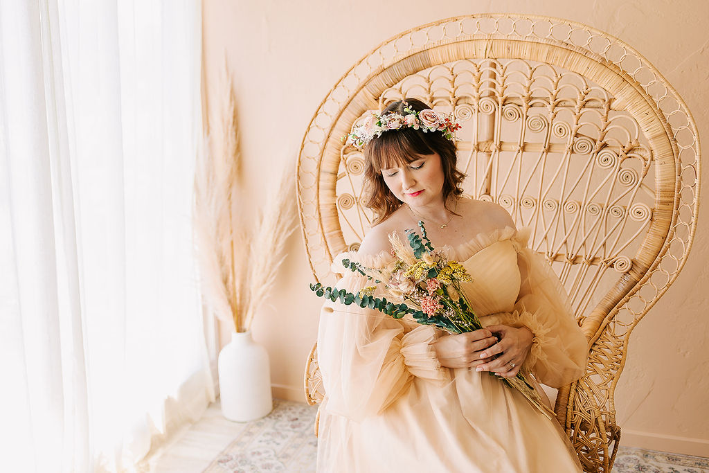 A mother to be sits in a tall wicker chair in a studio in a cream maternity gown holding some flowers after meeting a Midwife in Long Beach