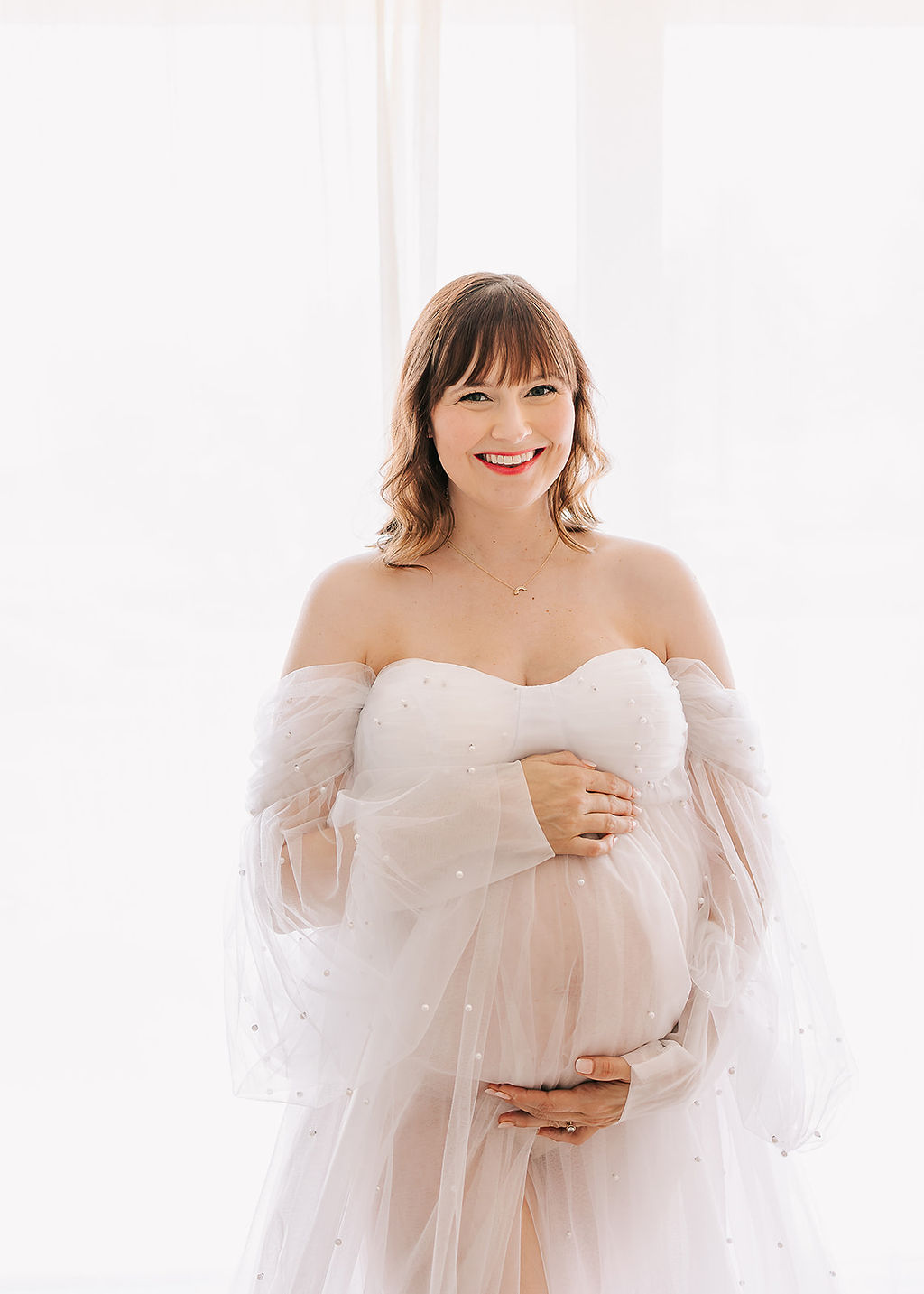 A happy mom to be smiles while holding her bump in a sheer maternity gown after meeting a Midwife in Long Beach