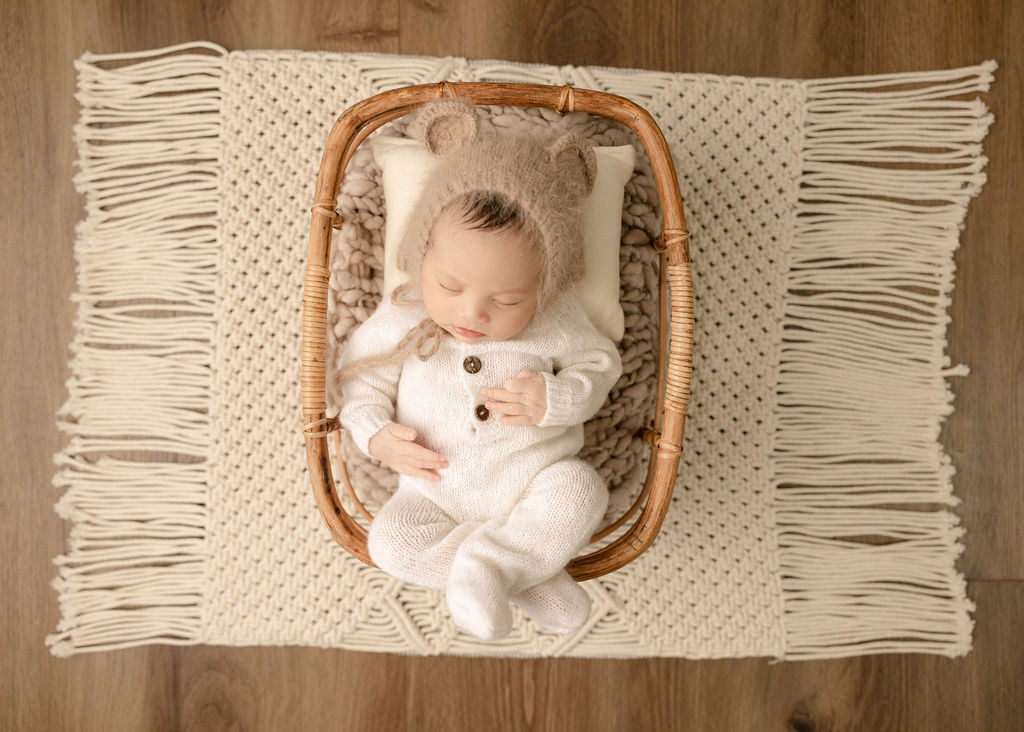 A newborn baby sleeps in a white knit onesie in a wicker basket