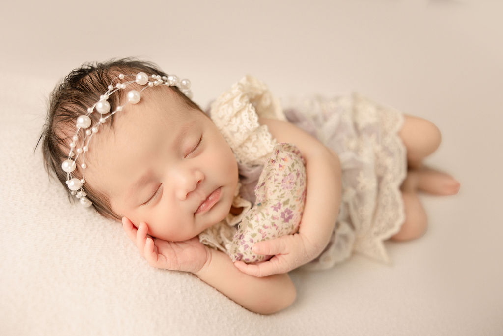 A sleeping newborn baby girl in a lace dress cuddles a small heart shaped pillow while on a white bed