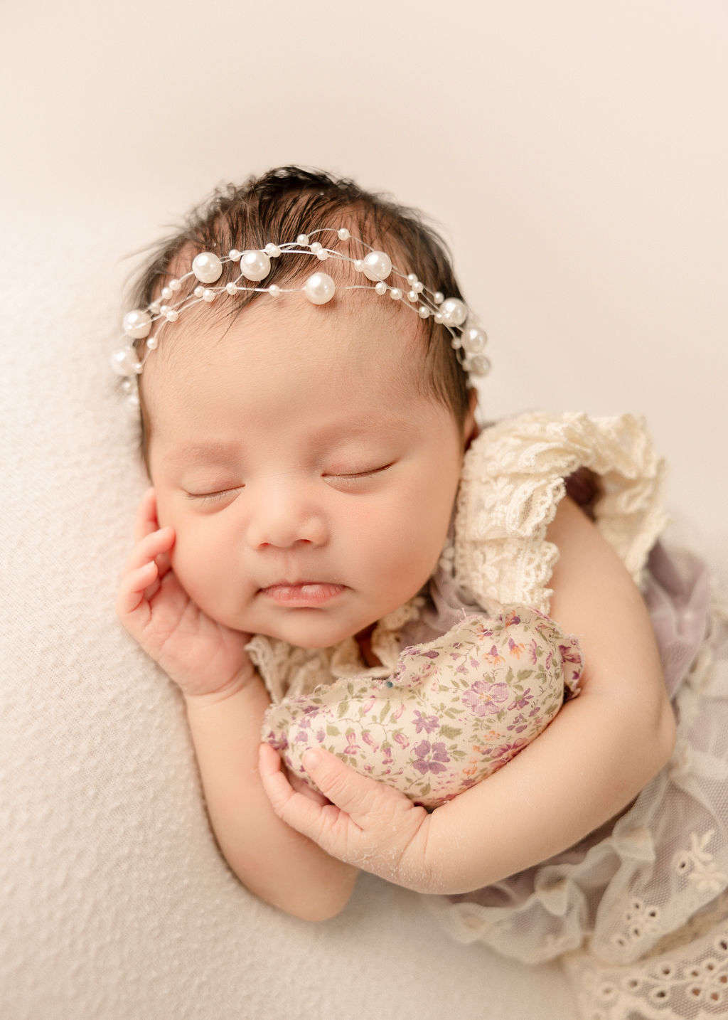 A sleeping newborn baby girl in a pearl headband and lace dress sleeps with a heart shaped pillow after meeting a nanny long beach