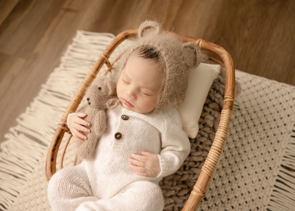 A newborn baby in a white knit onesie and bear bonnet cuddles a knit bear in a wicker basket after meeting a nanny long beach