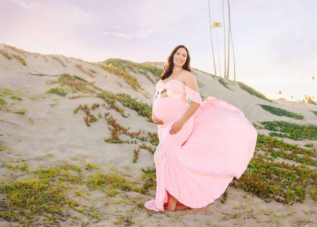 A happy mother to be stands on a windy beach at sunset in a pink maternity gown with her train blowing in the wind after visiting Baby Shower Venues in Long Beach