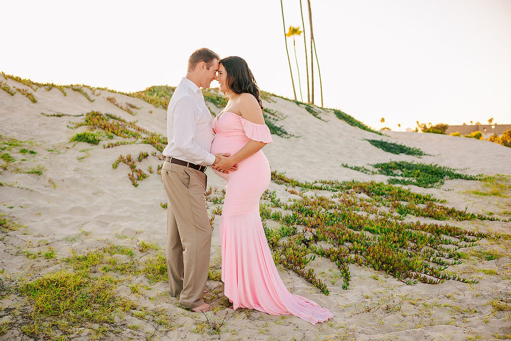 Expecting parents touch foreheads while smiling and holding the bump in a pink maternity gown