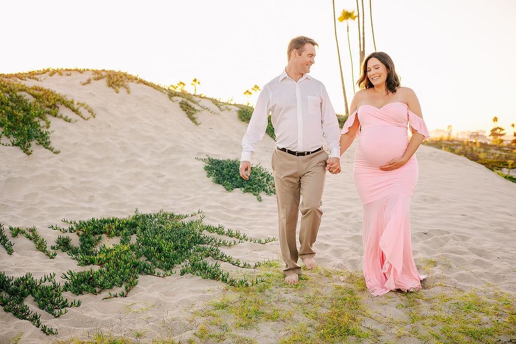 A happy expecting couple laughs while holding hands and walking on a beach dune at sunset after visiting Baby Shower Venues in Long Beach