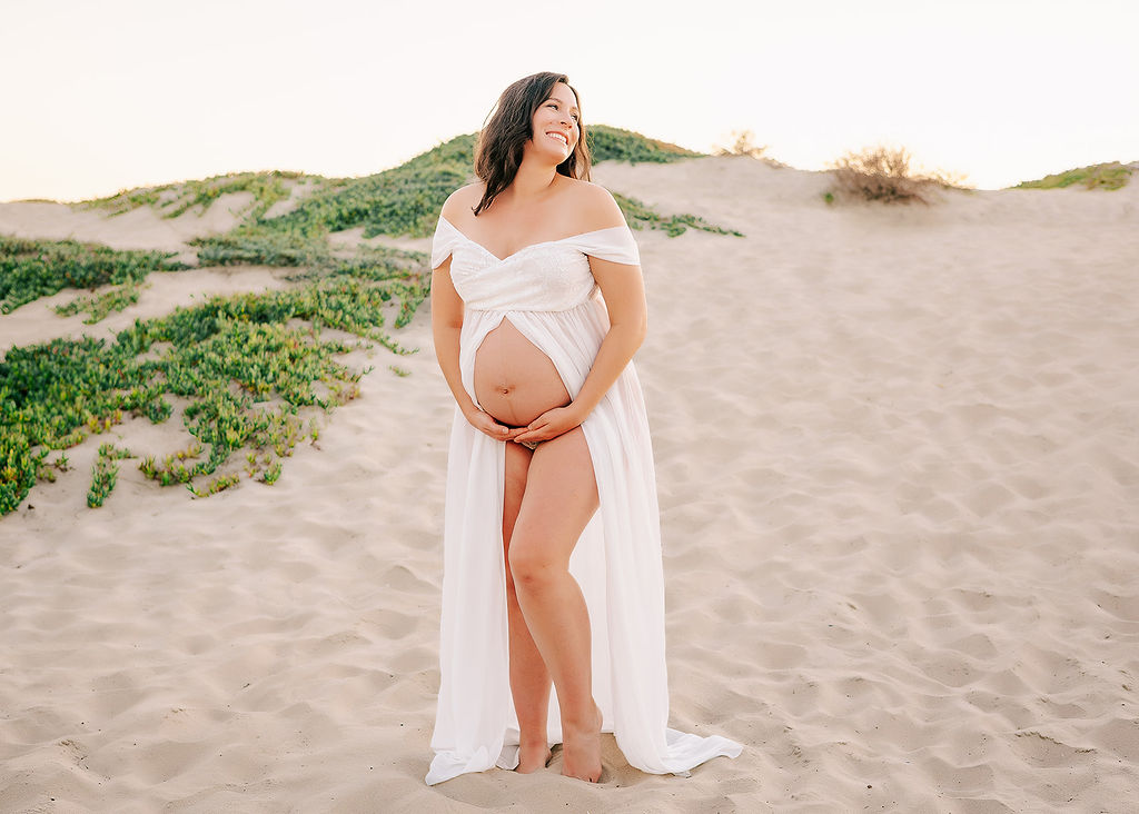 A laughing mother to be giggles while standing on a beach at sunset in an open white maternity gown before finding her Baby Shower Venues in Long Beach