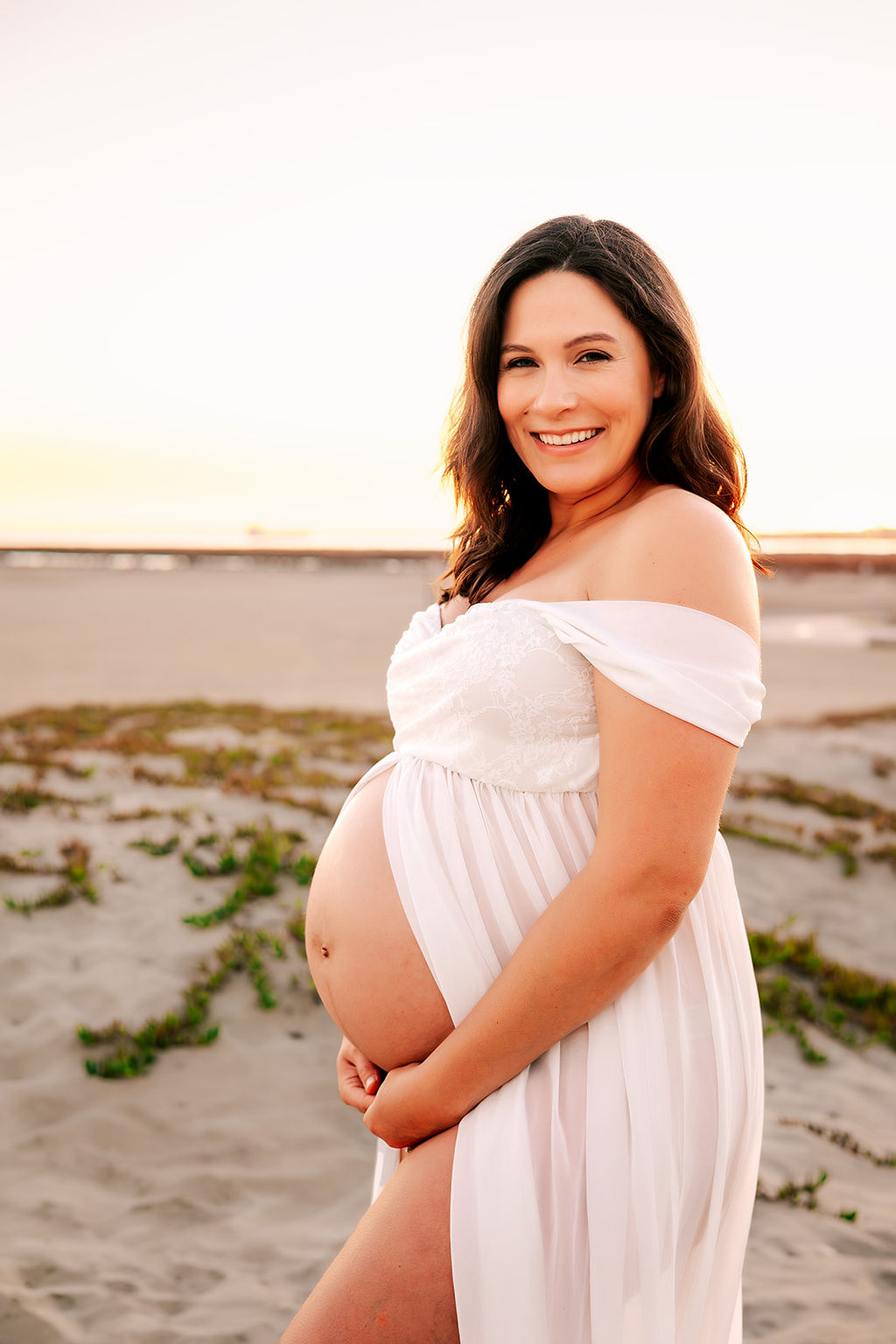 A happy mom to be in an open white maternity gown stands on a beach at sunset before visiting Baby Shower Venues in Long Beach