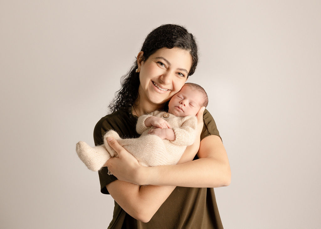 A happy new mother in a green shirt stands in a studio cradling her sleeping newborn baby in a white knit onesie