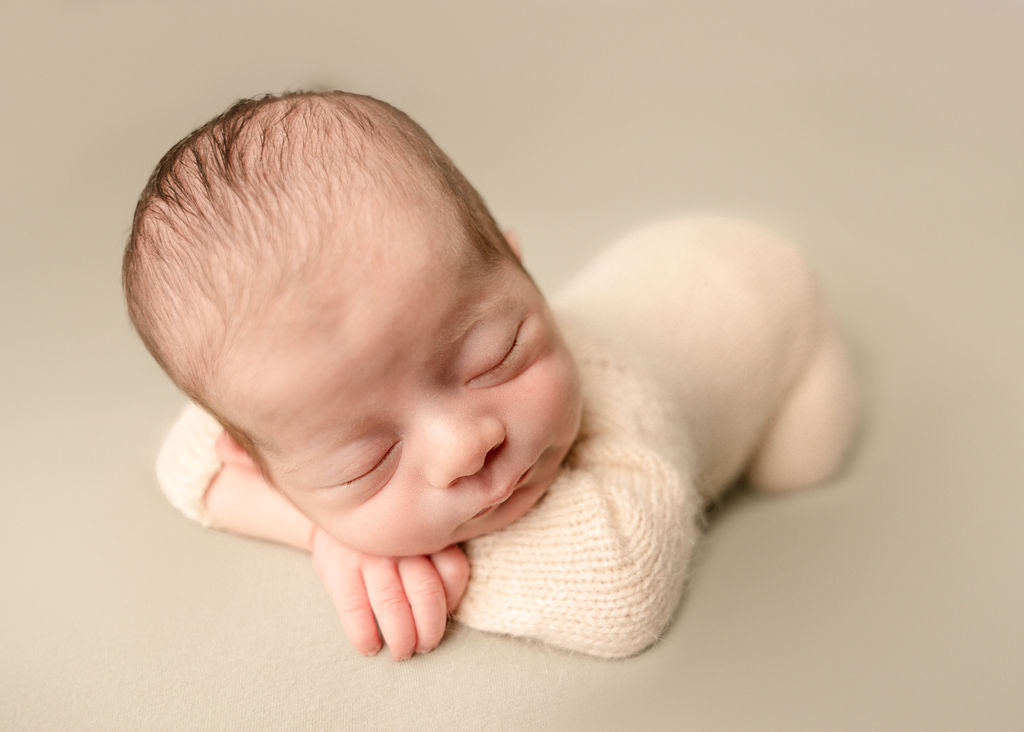 A newborn baby sleeps in froggy pose in a white knit onesie