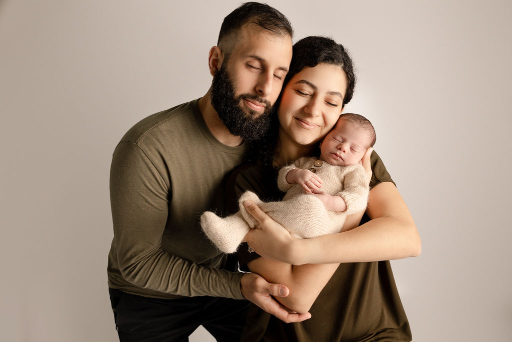 Happy new parents snuggle their sleeping newborn baby in a studio against their cheeks after visiting baby stores laguna beach