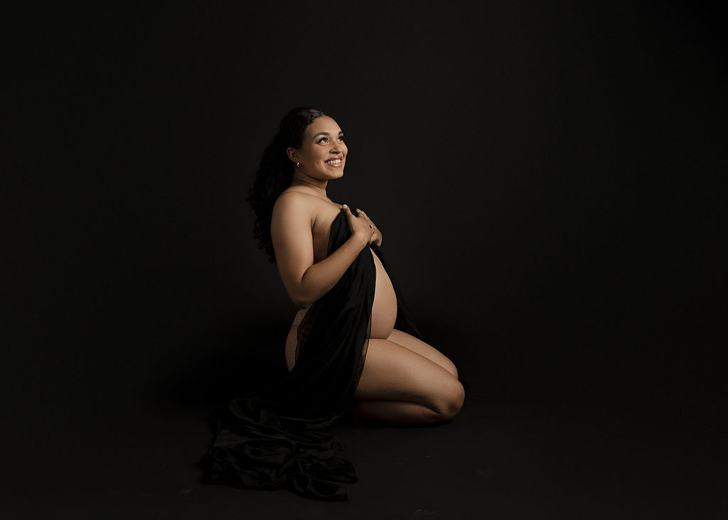 A smiling mother to be kneels in a dark studio while covering with a long black sheet