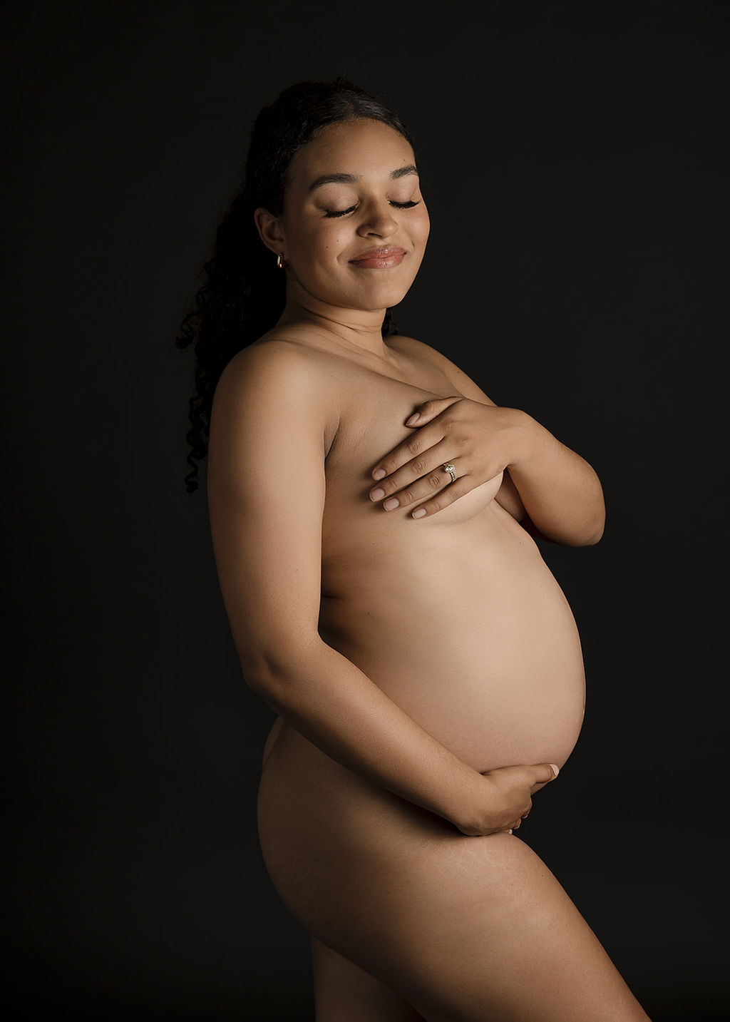 A mother to be smiles with eyes closed while covering her naked body with her hands