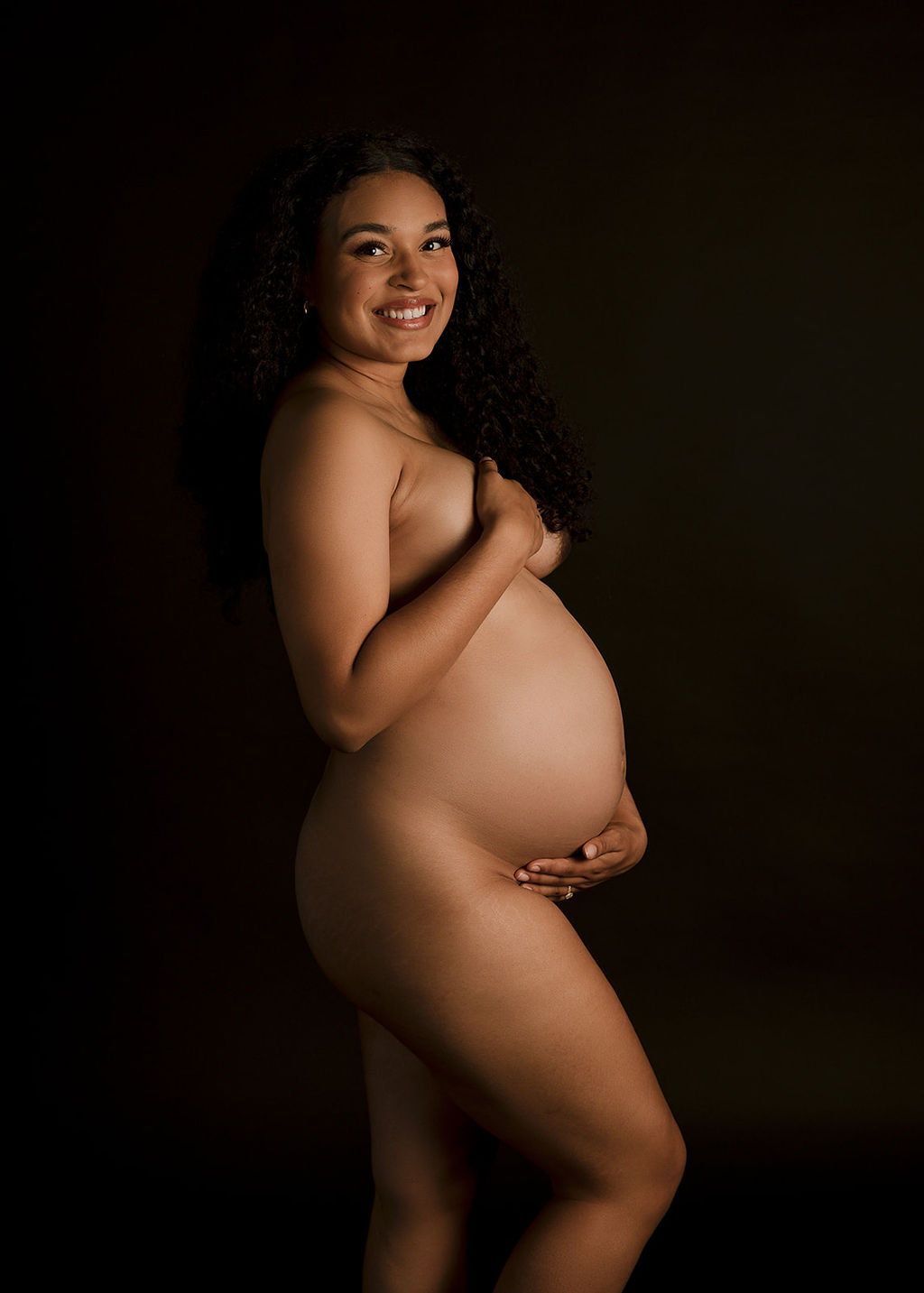 A naked pregnant woman smiles while standing in a studio with hands covering herself after meeting Boise Doulas