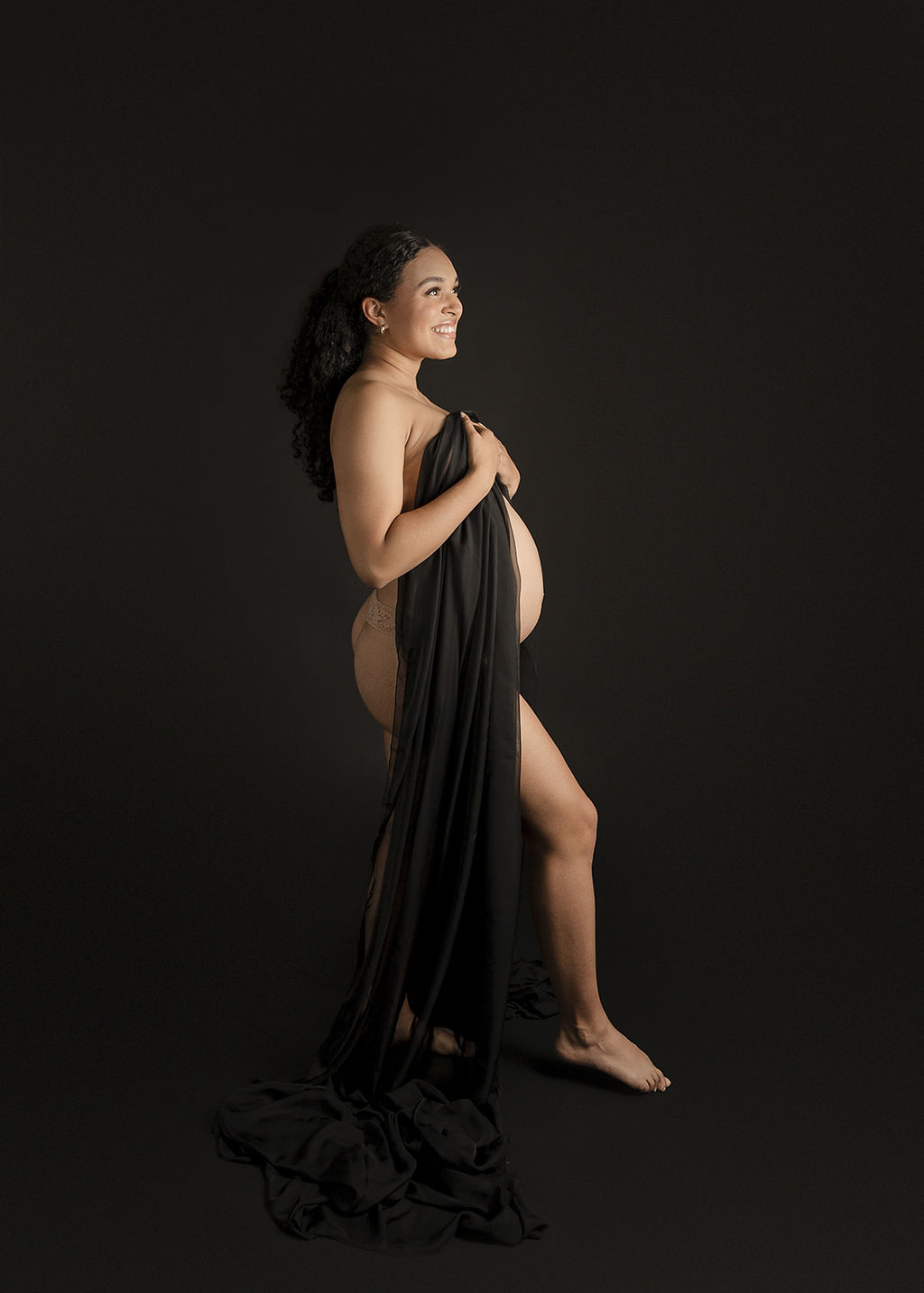 A smiling mom to be stands in a studio covering with a long black sheet in her underwear after meeting with Boise Doulas