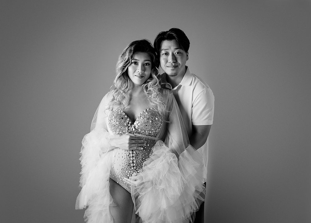 A happy pregnant couple hugs while standing in a studio smiling