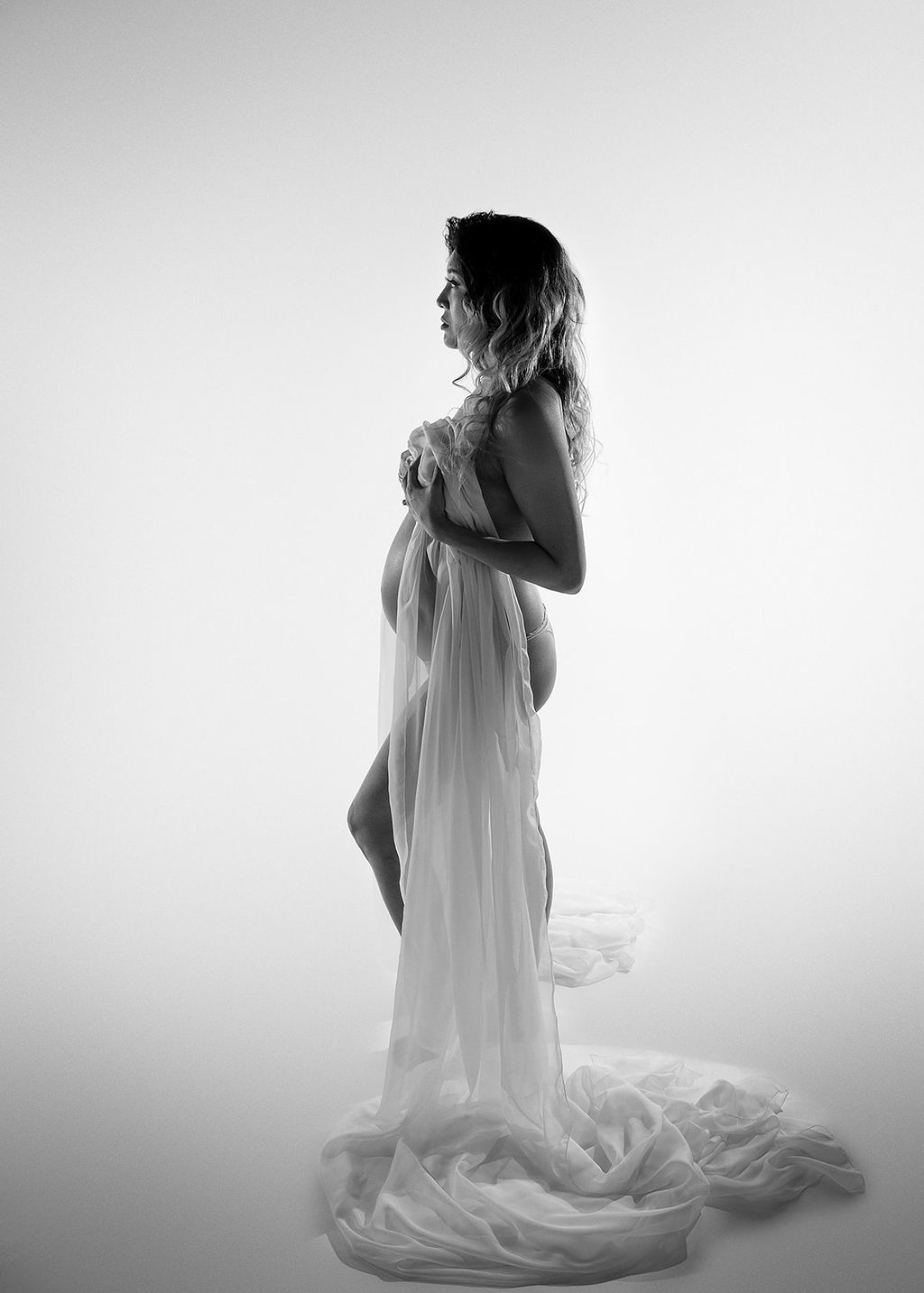 A pregnant woman covers with a long sheer sheet while standing in a studio in underwear after meeting Boise Midwives