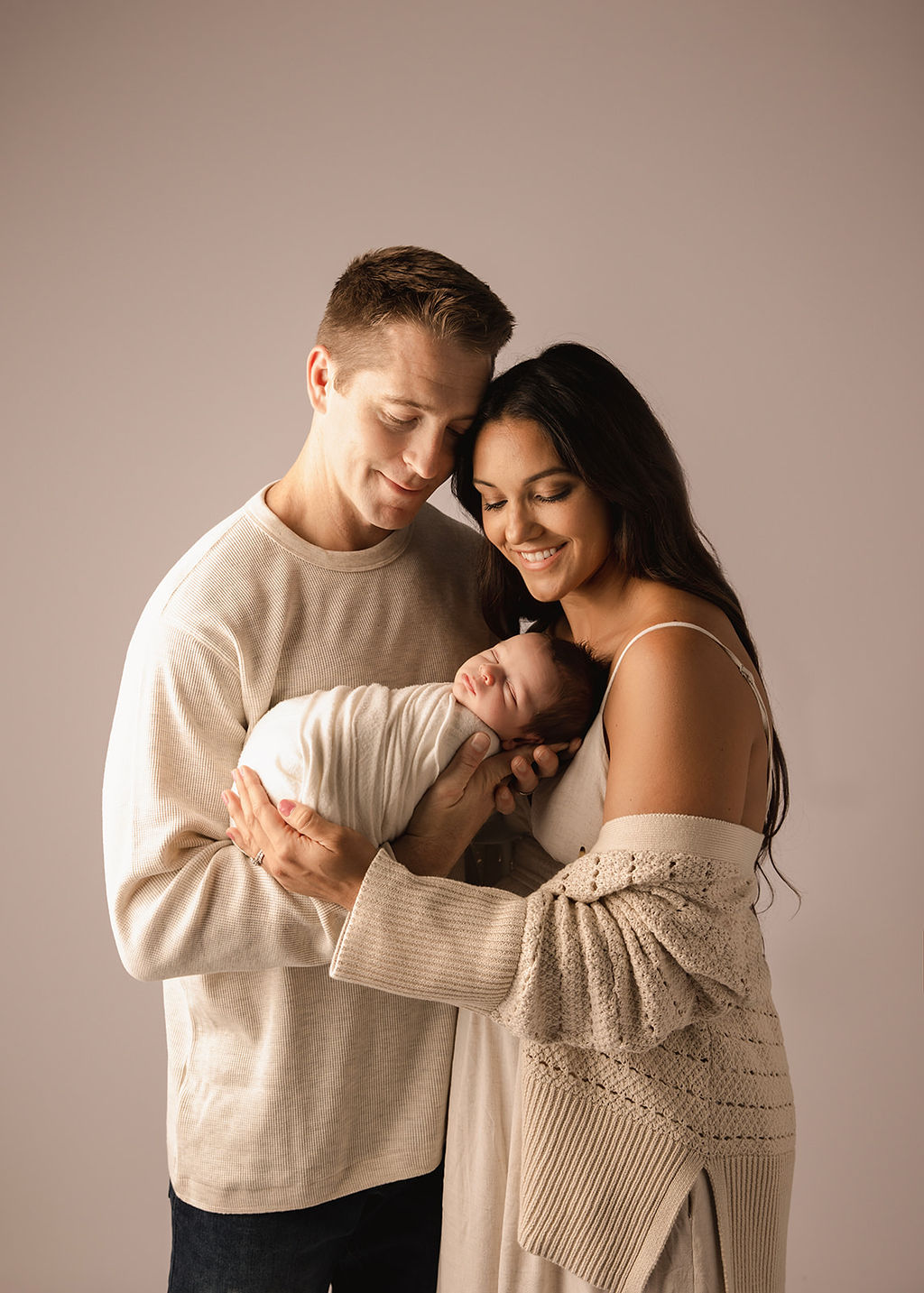 Happy new parents snuggle while standing in a studio with their sleeping newborn baby between them