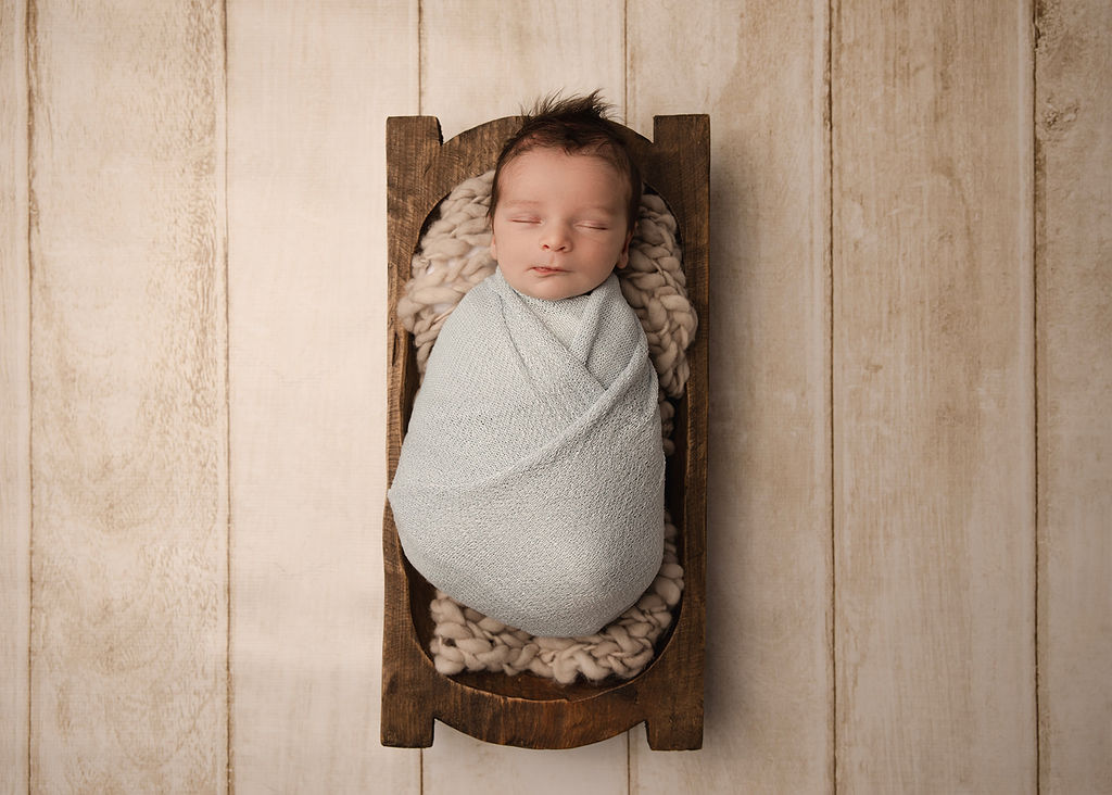 A newborn baby sleeps in a wooden bowl in a blue swaddle in a studio