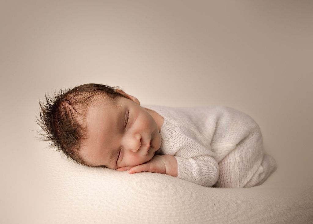 A newborn baby sleeps on its tummy on a bed in a studio in a white knit onesie before visiting a Daycare Long Beach