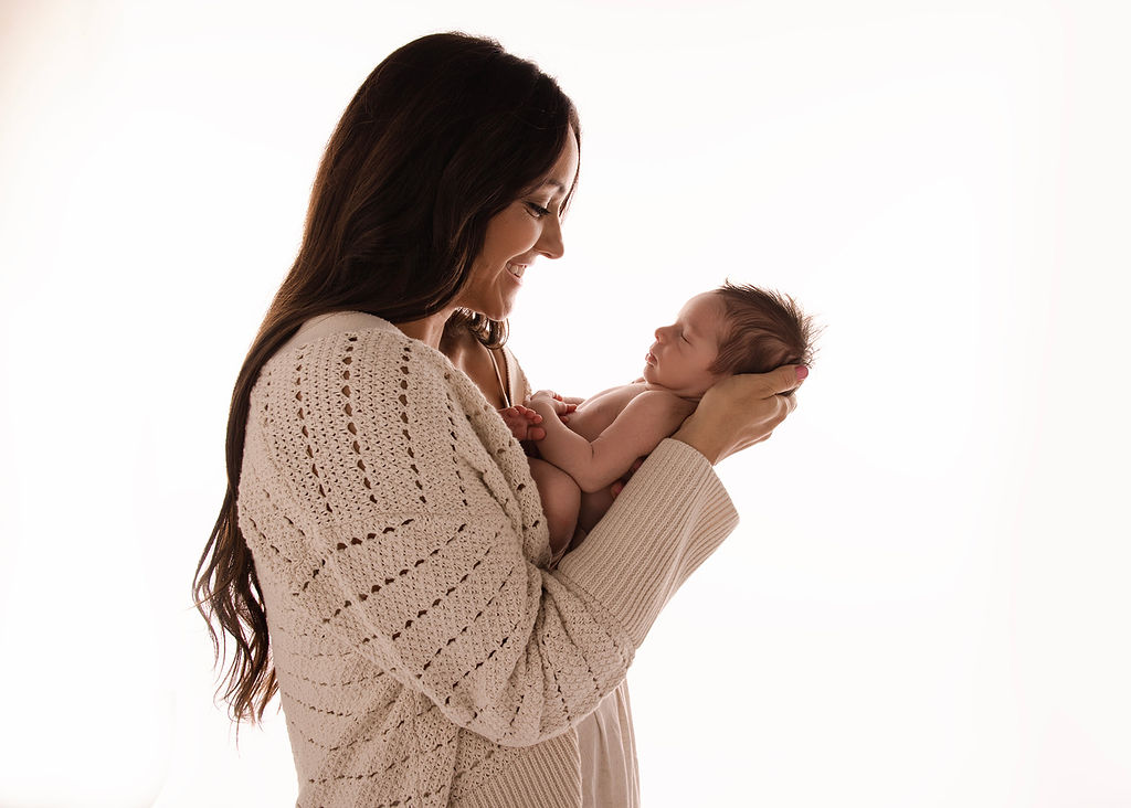 A happy new mom in a knit sweater smiles down at her newborn baby in her hands while standing in a window before finding a Daycare Long Beach