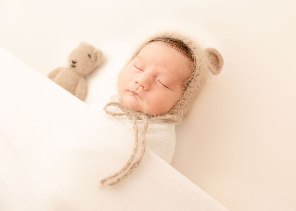 A newborn abby sleeps in a knit teddy bonnet with a matching bear on a bed under a blanket