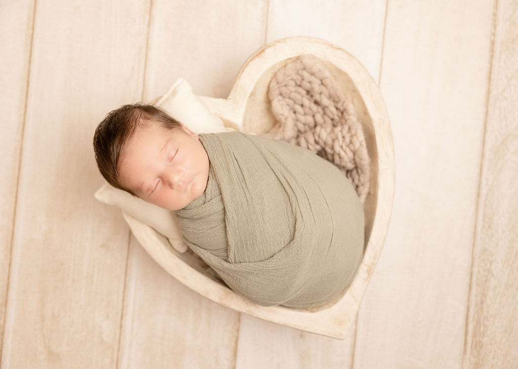 A newborn baby in a tight green swaddle sleeps in a wooden heart shaped bowl