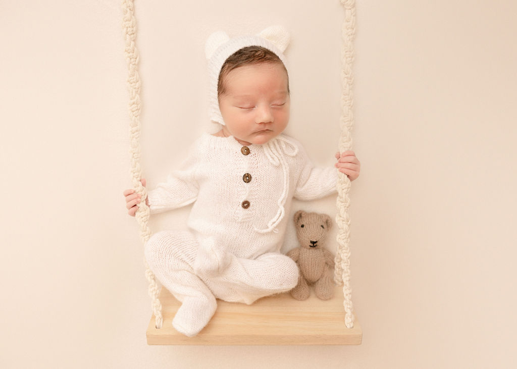 A newborn baby in a white knit onesie sits on a swing while sleeping with a brown teddy bear