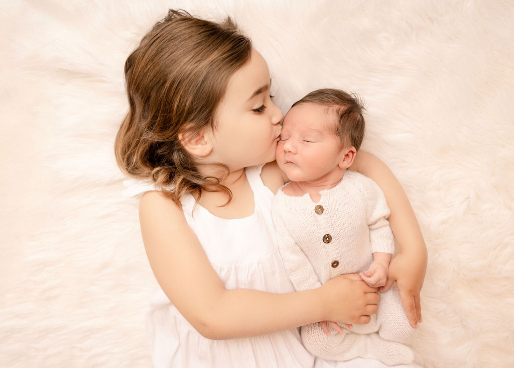 A toddler girl in a white dress kisses the cheek of her newborn baby brother while using diaper service long beach