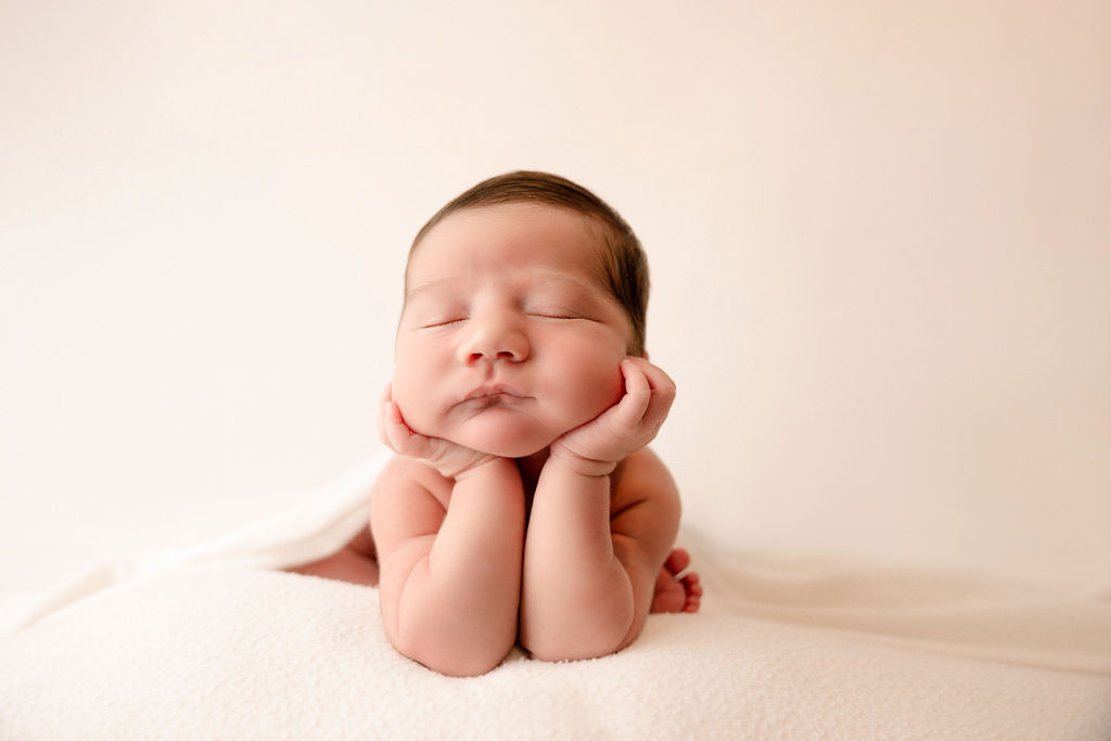 A newborn baby rests its head on its hands while sitting on a bed in a studio after using diaper service long beach