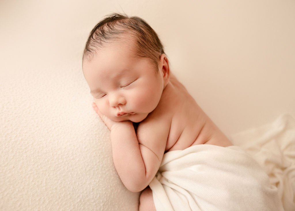 A newborn baby sleeps on its hand under a white blanket in a studio