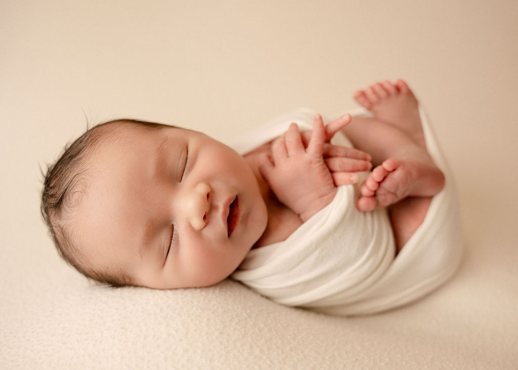 A sleeping newborn baby in a white swaddle with hands and legs poking out on a white bed after mommy and me classes long beach