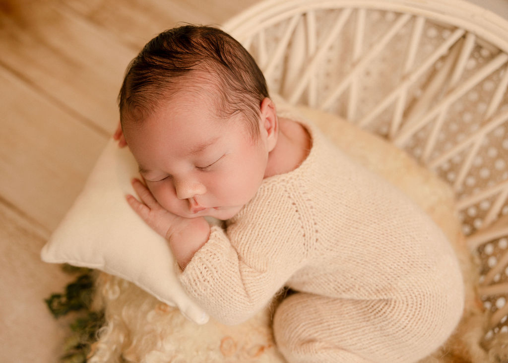 A newborn baby sleeps in a white knit onesie in a wicker basket on a pillow after some mommy and me classes long beach