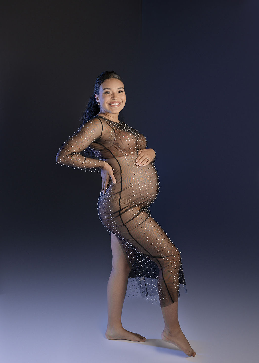 A happy mother to be smiles while resting a hand on her bump and standing in a studio in a sheer maternity gown after a prenatal massage long beach