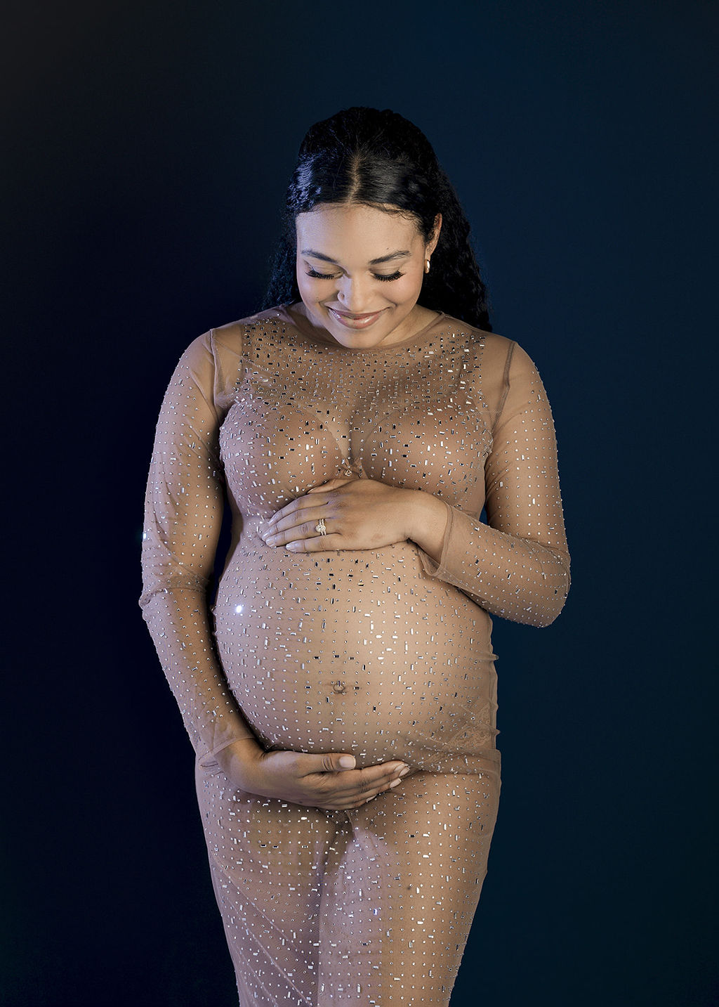 A happy mom to be stands in a studio in a sheer maternity gown