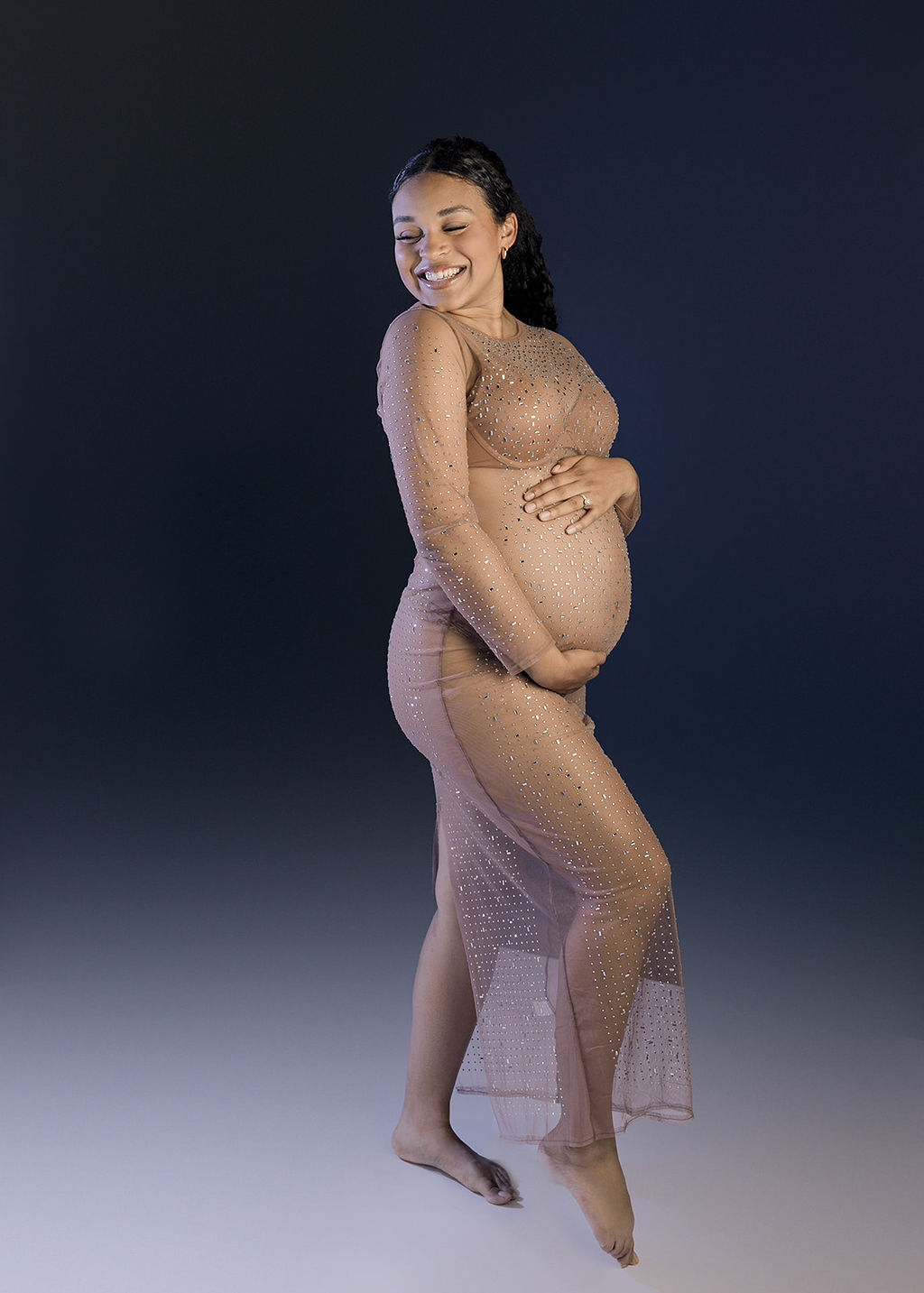 A happy mother to be smiles over her shoulder while holding her bump in a nude maternity gown with rhinestones after a prenatal massage long beach