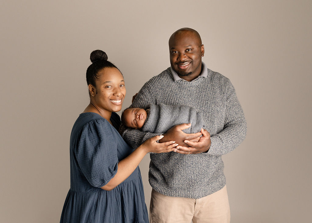 Happy new parents stand in a studio cradling their sleeping newborn baby before visiting baby stores in Boise