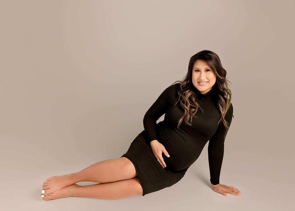 A smiling mom to be sits across the floor in a studio holding her bump and smiling