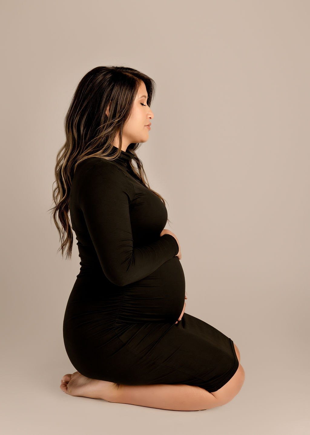 A pregnant woman in a black dress kneels on the floor of a studio with hands on her bump