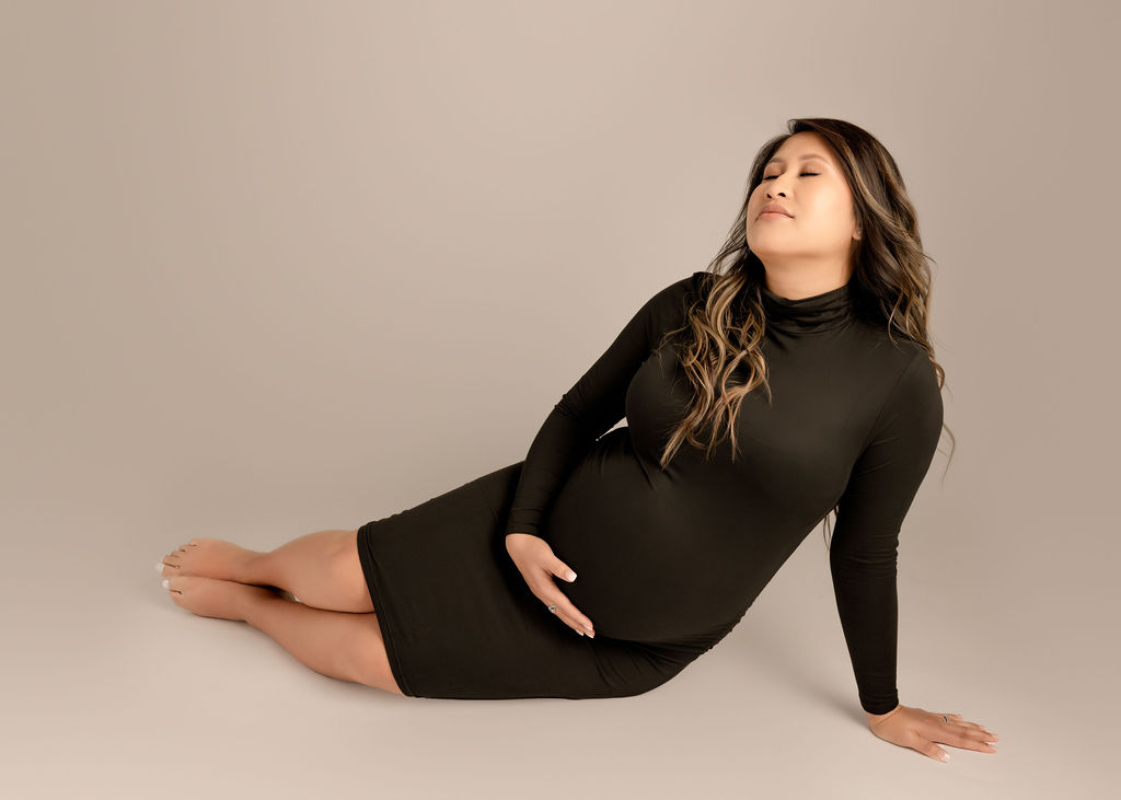 A happy mother to be sits across the floor of a studio in a black maternity gown with a hand on her bump after visiting a birth center in boise