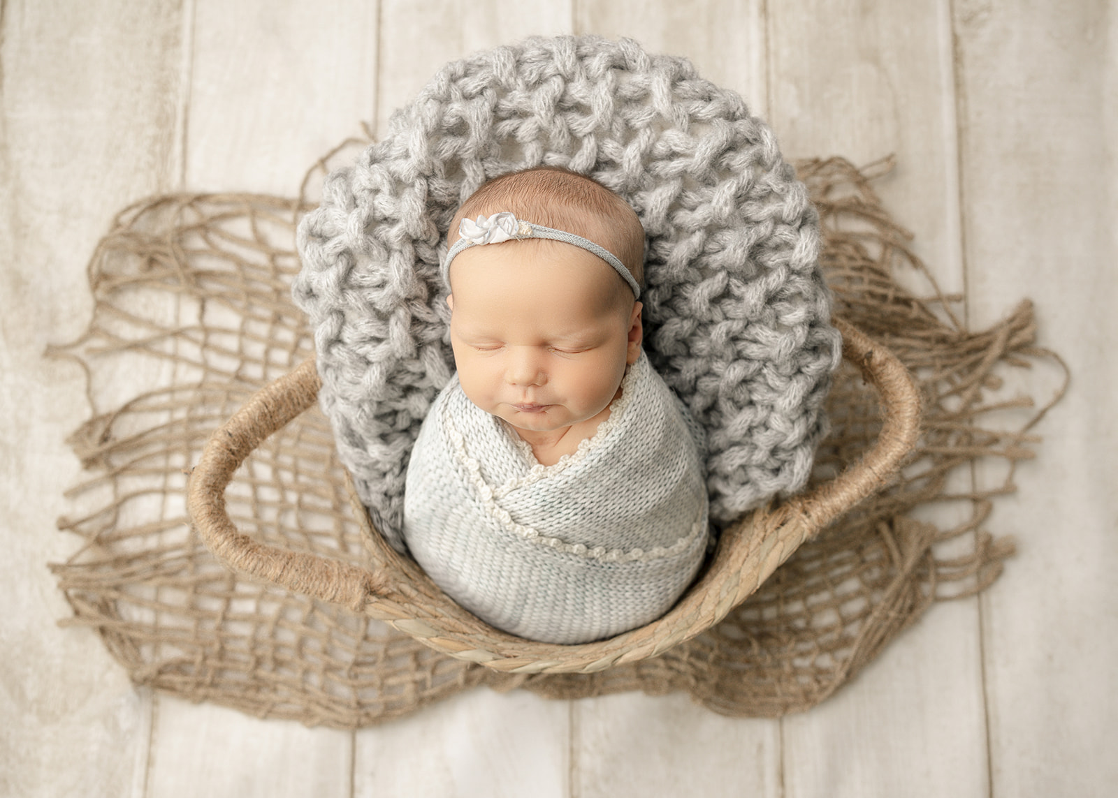 A newborn abby sleeps in a grey swaddle and matching headband in a woven basket in a studio