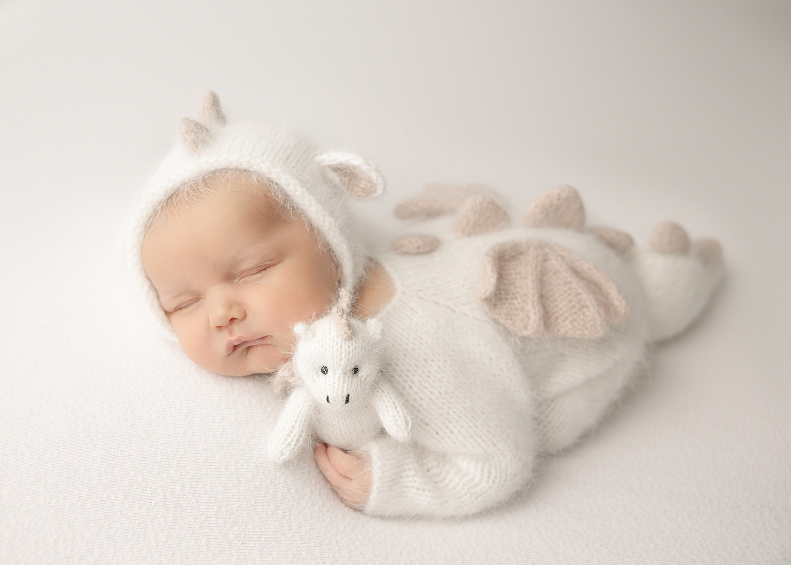 A newborn baby sleeps in a knit white dragon onesie while holding a matching stuffy after meeting a lactation consultant in Boise